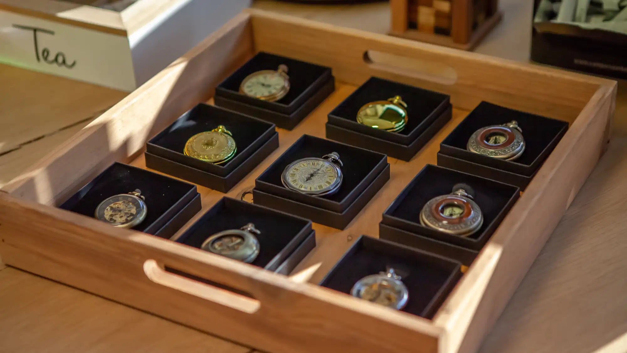 Une collection de neuf montres de poche ornées, chacune nichée dans une boîte doublée de velours noir au sein d'un plateau en bois clair.