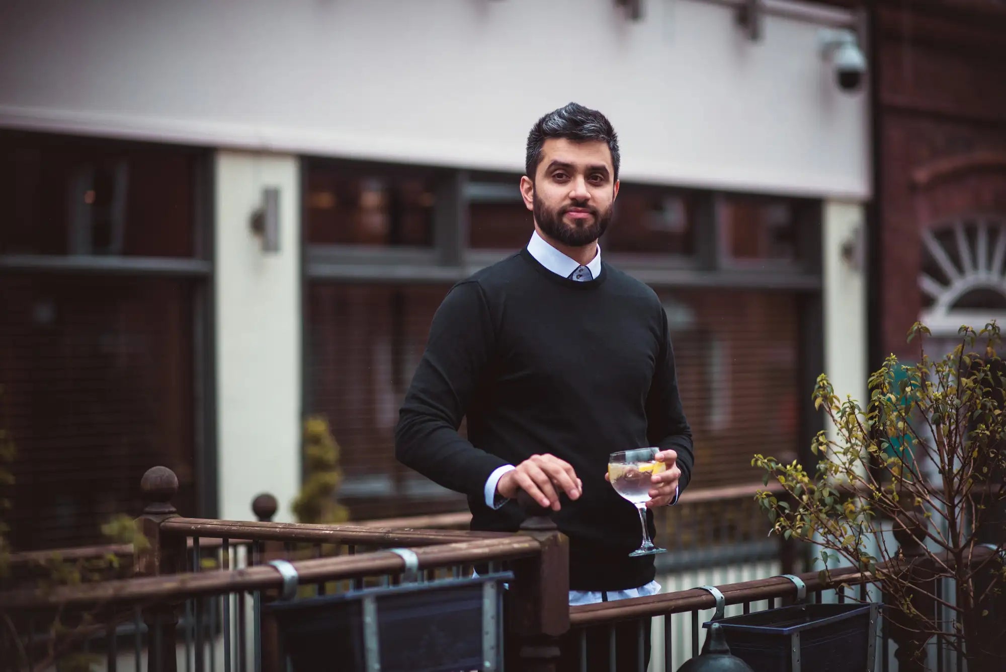 Un homme portant un pull noir et une chemise à col clair bleu pâle tient un verre à pied transparent contenant une boisson ambrée garnie d'une torsade de citron.