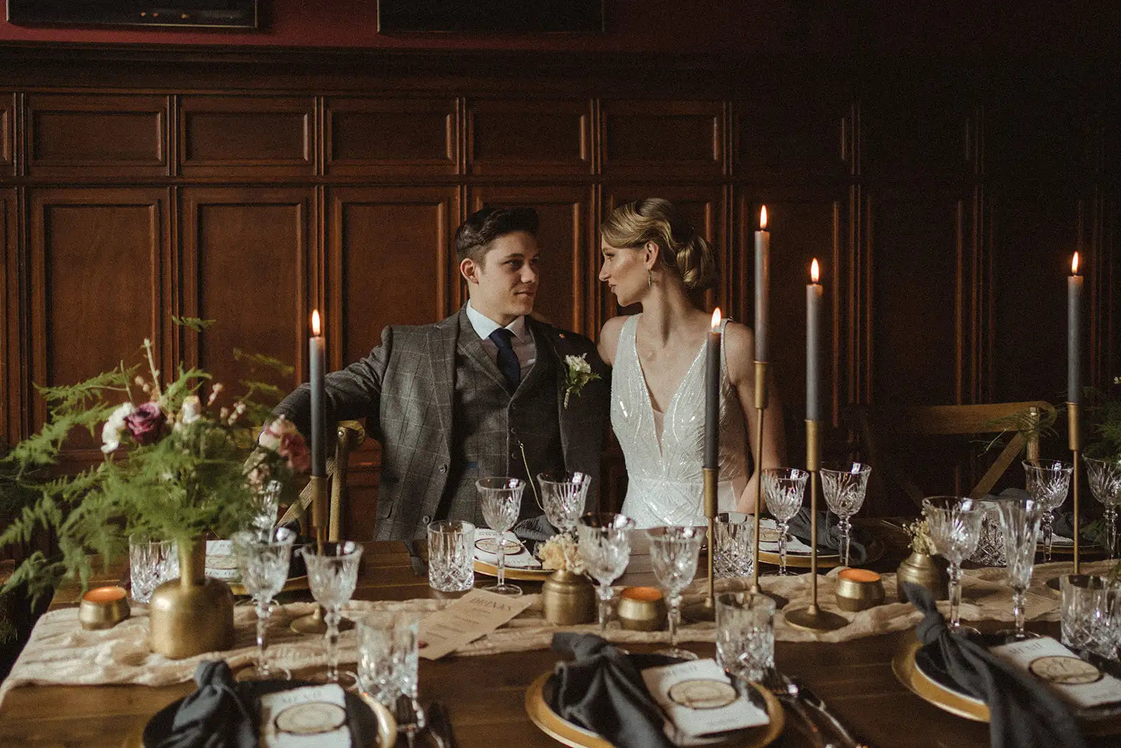 Verres à vin en cristal élégants avec des motifs gravés complexes, posés sur une table en bois sombre aux côtés de chandeliers aux tons dorés et de serviettes noires.